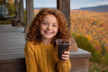 Smiling girl in a yellow sweater on a porch step holding up a cup of potting mix with an apple twig cutting, West Virginia hills in fall color behind her