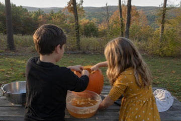 Two children at a backyard picnic table near Ozark woods scooping seeds from a pumpkin into a bowl of water