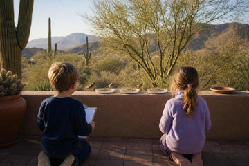 Children watching small birds feed from three dishes on a desert patio wall with saguaro cacti and mountains behind
