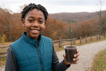 A smiling boy in a quilted vest holds up a small plant in a clear cup on a gravel path, with a wooden fence and fall-colored trees behind him