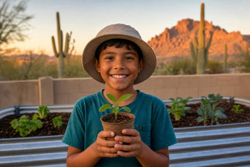 A smiling boy in a teal T-shirt and sun hat holds up a potted seedling in front of raised garden beds, with saguaro cacti and desert mountains under a sunset sky behind him