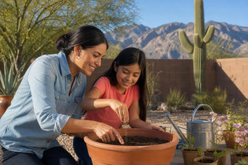 A smiling mother and daughter with dark hair kneel over a large terracotta pot, planting seeds together in a desert backyard with a watering can, small potted plants, a cactus and mountains behind them
