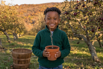 A smiling boy in a dark green zip fleece faces the camera in an apple orchard, holding up a terracotta pot with a thin young twig, with red-apple trees, a wooden basket and autumn hills behind him