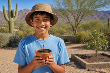 A smiling boy in a tan bucket hat and light blue T-shirt faces the camera holding a terracotta pot with a thin twig cutting, in a desert yard with cactus, trees, a raised bed and mountains behind him