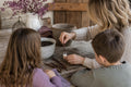 A parent and two children at a farmhouse table dipping a bare, leafless plum tree cutting into rooting powder
